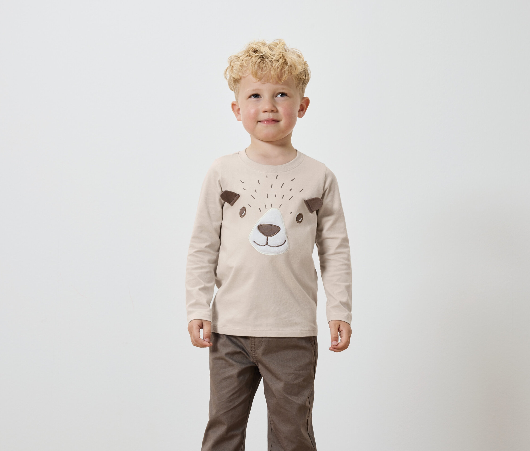 Un garçon aux cheveux blonds bouclés porte un t-shirt beige à manches longues avec des détails marron représentant un visage d'ours et un pantalon marron.