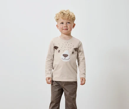 Un garçon aux cheveux blonds bouclés porte un t-shirt beige à manches longues avec des détails marron représentant un visage d'ours et un pantalon marron.