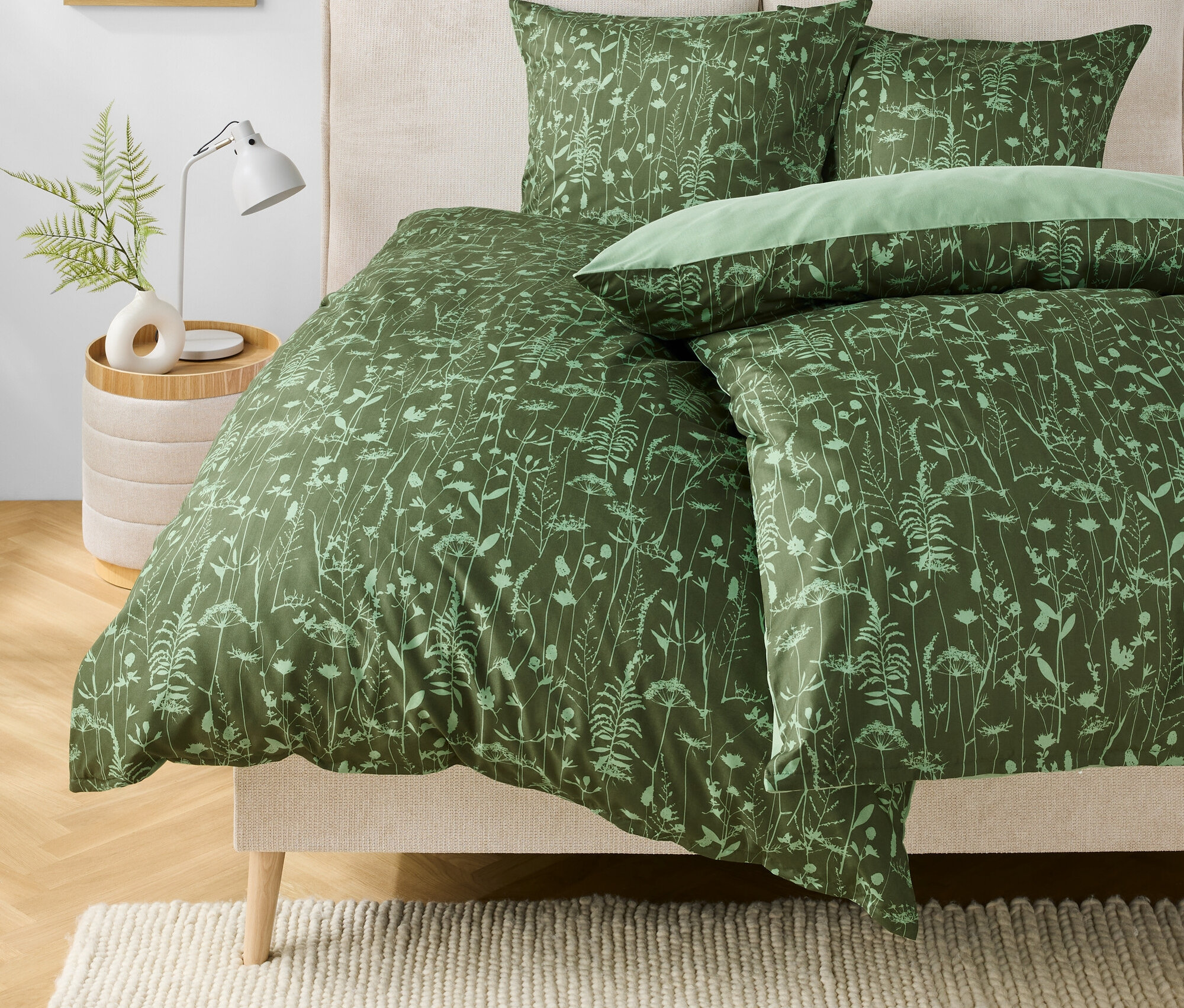 Linge de lit vert à motifs floraux sur un lit. Une lampe et un vase avec une plante sont posés sur la table de chevet.