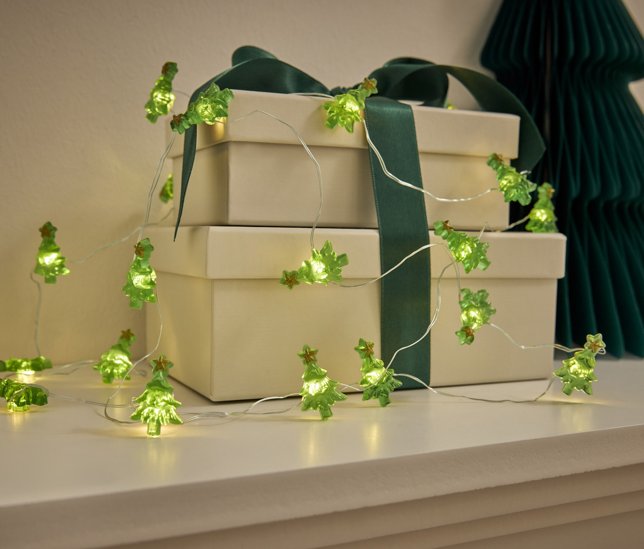 Guirlande lumineuse verte en forme de sapin de Noël sur une commode blanche avec des cadeaux et un arbre en papier.