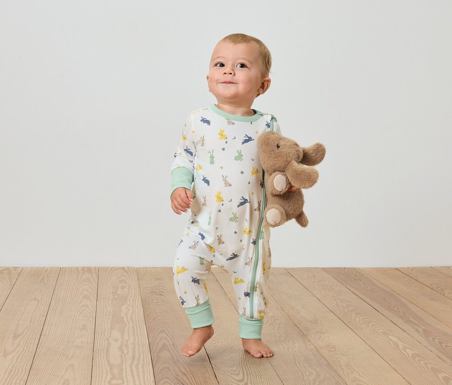 Un bébé se tient sur un plancher en bois et tient un éléphant en peluche.