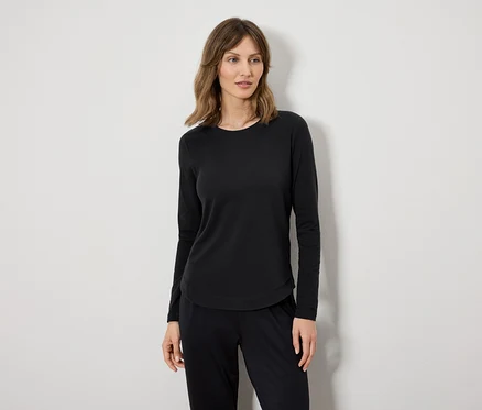 Une femme portant un t-shirt à manches longues noir et un pantalon noir se tient devant un mur blanc.