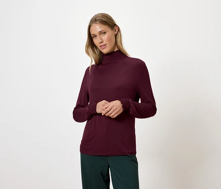Une femme blonde porte un pull à col roulé bordeaux et un pantalon vert.