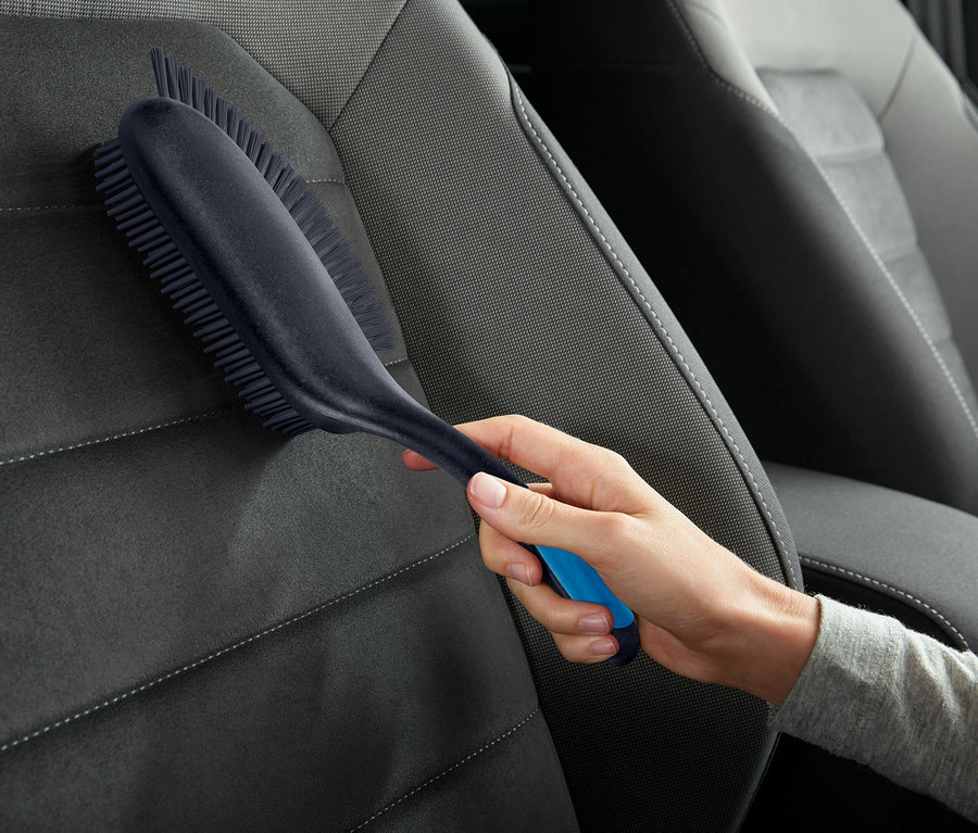 Une main tient une brosse avec des accents bleus et nettoie un siège auto gris.