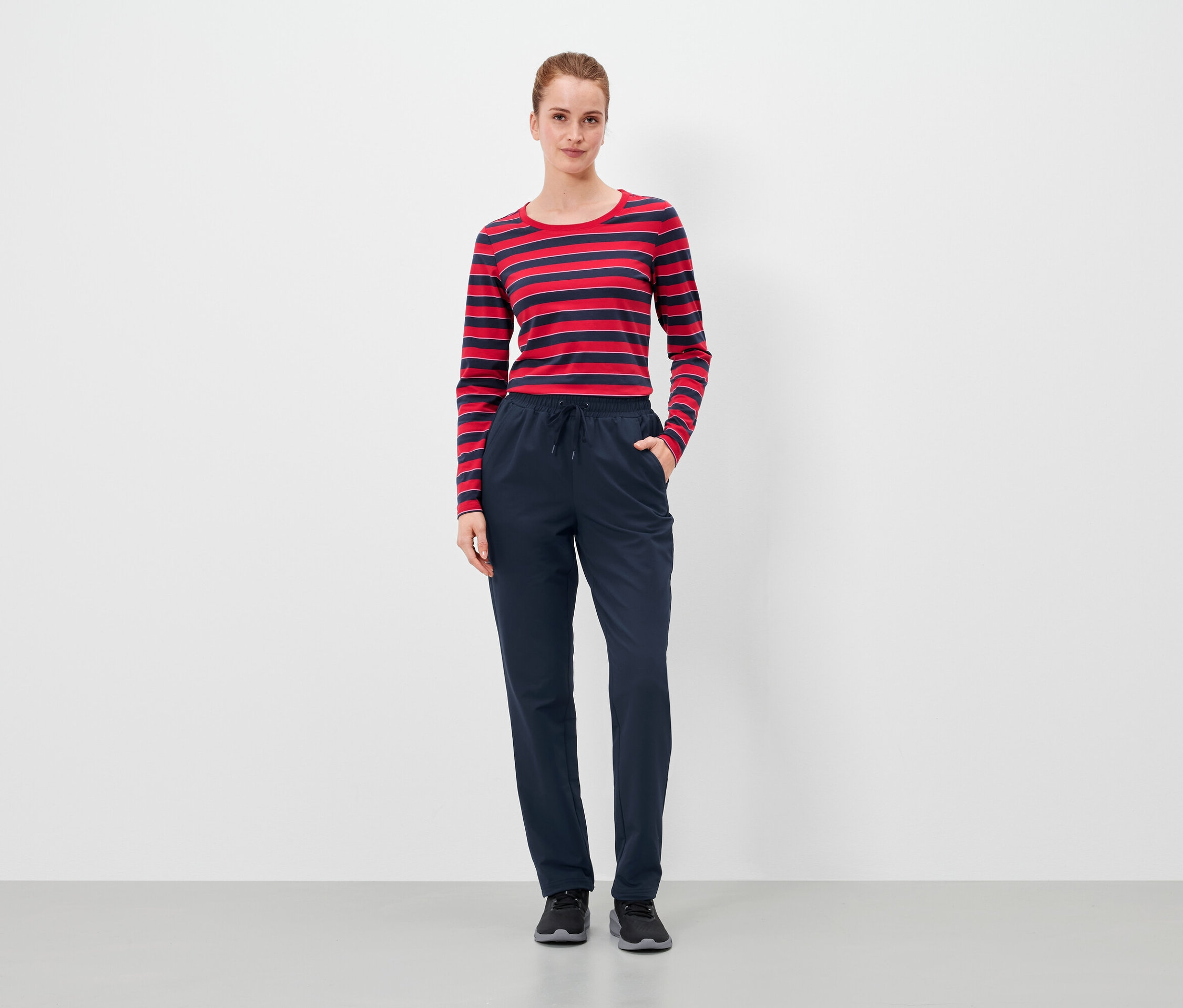 Une femme se tient debout, les mains dans les poches d'un pantalon bleu marine, et porte un t-shirt à rayures rouges et bleues.