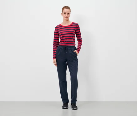 Une femme se tient debout, les mains dans les poches d'un pantalon bleu marine, et porte un t-shirt à rayures rouges et bleues.