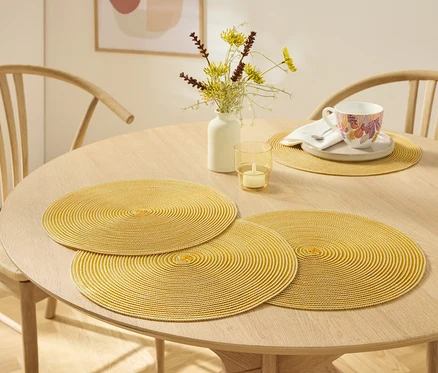Sur une table en bois clair se trouvent trois sets de table ronds jaunes, un vase avec des fleurs, une bougie et une tasse avec une soucoupe.