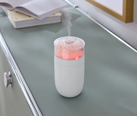 Sur une commode grise se trouve un humidificateur blanc avec des pierres roses et de la vapeur.