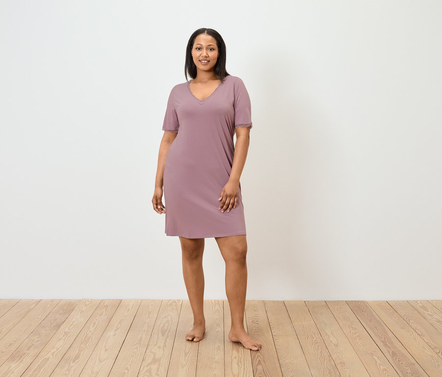 Une femme se tient pieds nus et porte une courte chemise de nuit mauve.