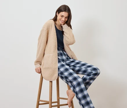 Une femme est assise sur un tabouret, portant un manteau beige duveteux et un pantalon de pyjama à carreaux bleus.
