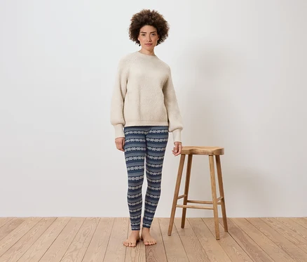 Une femme se tient devant un mur blanc à côté d'un tabouret en bois. Elle porte un pull crème et un legging bleu à motifs.