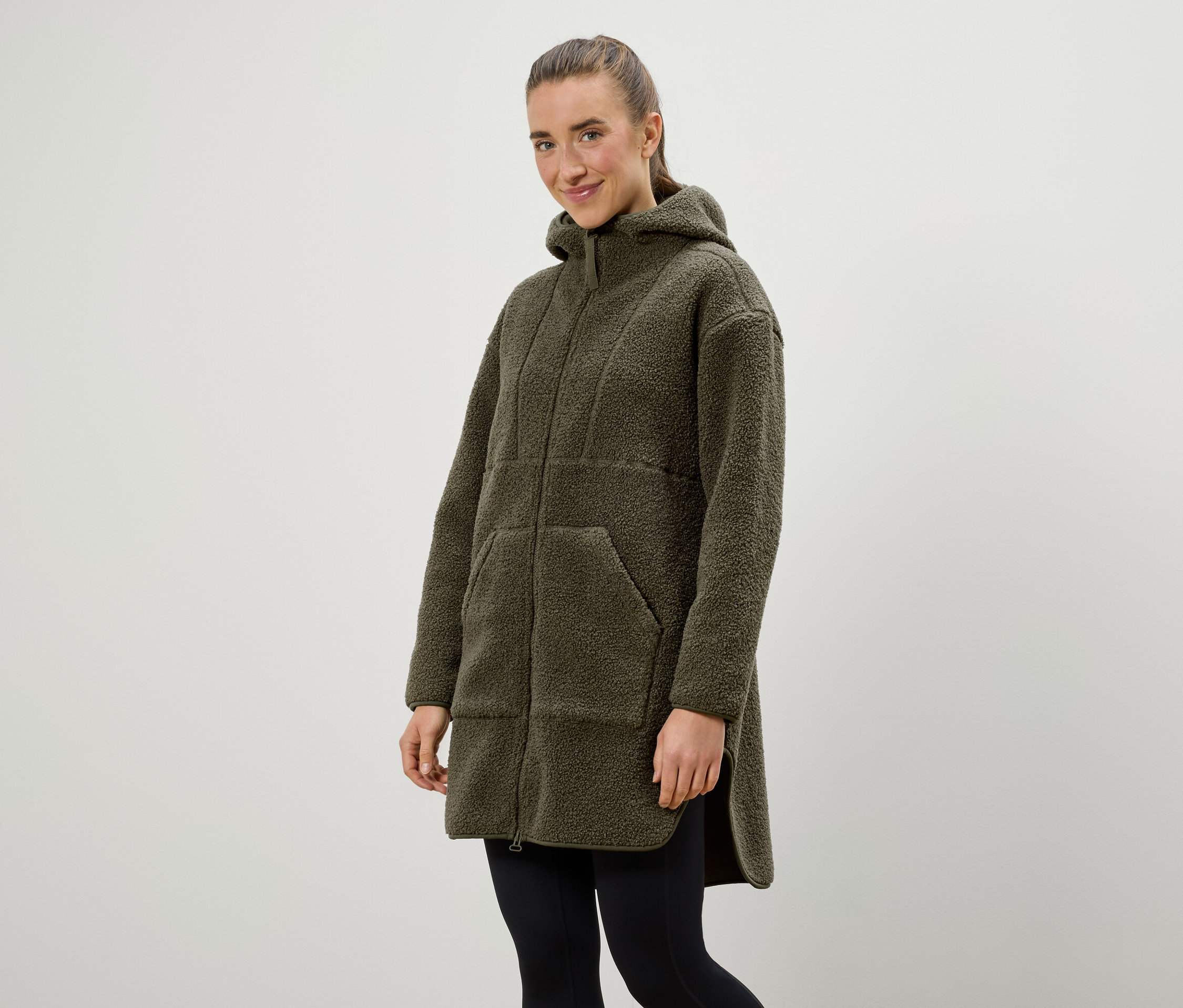 Une femme porte un manteau à capuche vert olive et un legging noir.