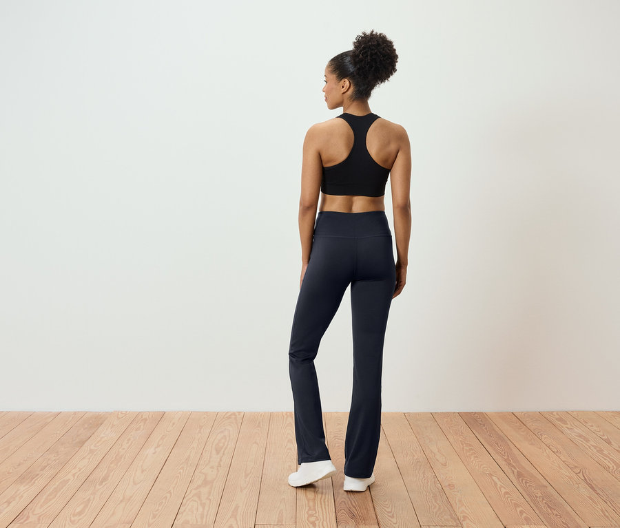 Vue de dos d'une femme aux cheveux relevés portant un haut de sport noir et un pantalon bleu marine, debout sur un plancher en bois.