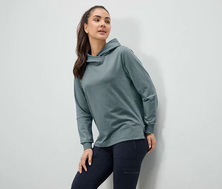 Une femme pose en sweat à capuche bleu-vert et pantalon bleu foncé devant un mur blanc.