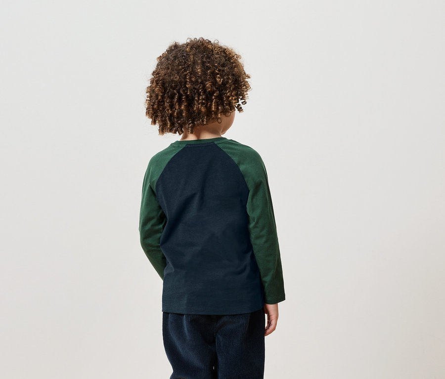 Un garçon aux cheveux bouclés se tient dos à la caméra, portant un t-shirt à manches longues vert et un pantalon en velours côtelé bleu marine.