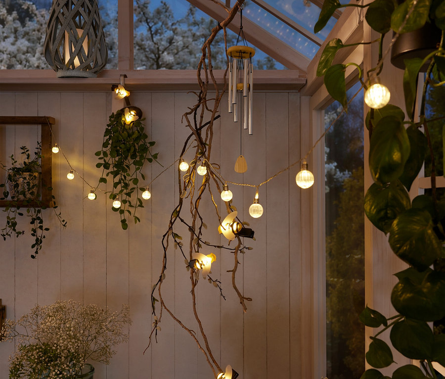 Intérieur d'une véranda avec une lanterne grise, une guirlande lumineuse avec des boules et des abeilles, un carillon éolien et des plantes.