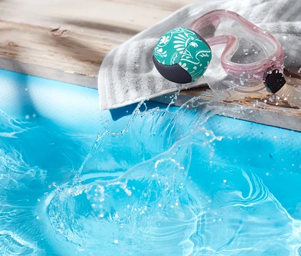Un ballon vert à motifs blancs, des lunettes de natation et une serviette reposent sur une terrasse en bois à côté d'une piscine où l'eau éclabousse.