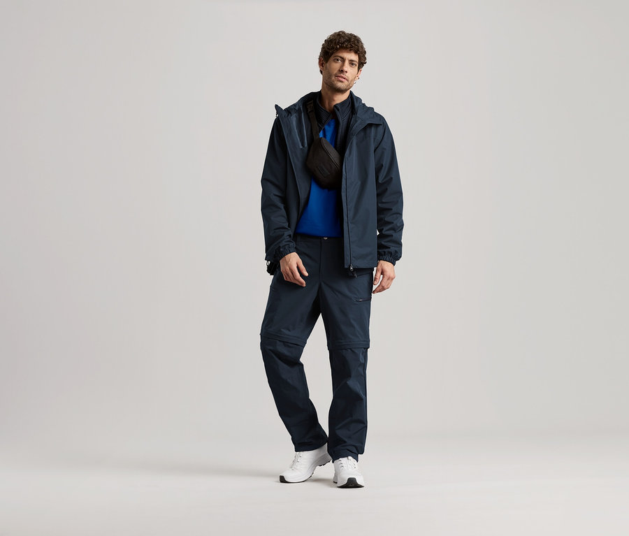 Un homme debout en veste bleu marine, pantalon et baskets blanches, avec un sac bandoulière noir.