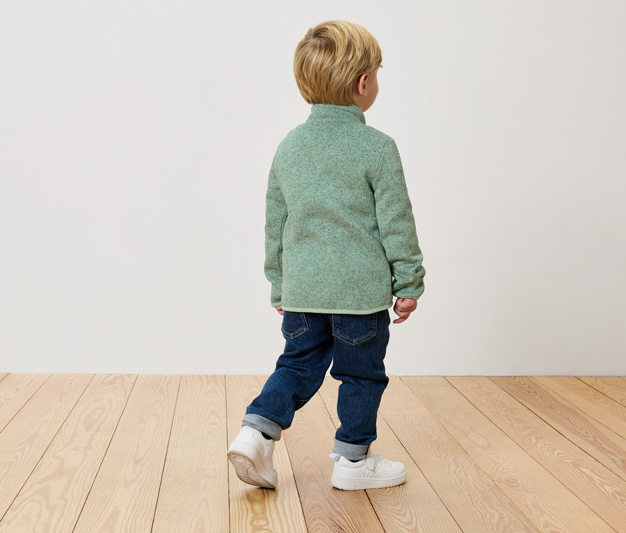 Un enfant marche dos à la caméra dans un pull en polaire vert, un jean et des baskets blanches sur un plancher en bois.