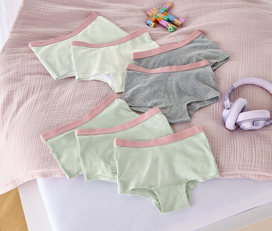 Sur un lit sont disposés sept culottes pour filles rose fané, un jouet et un casque audio.