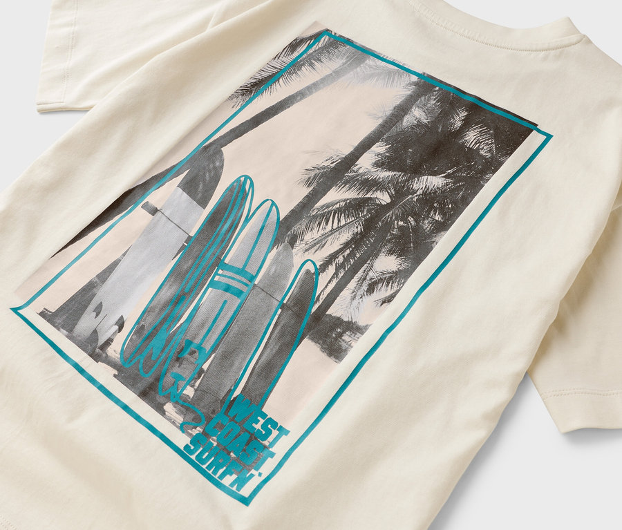 Sur un t-shirt crème, on voit un rectangle imprimé avec des palmiers et des planches de surf.