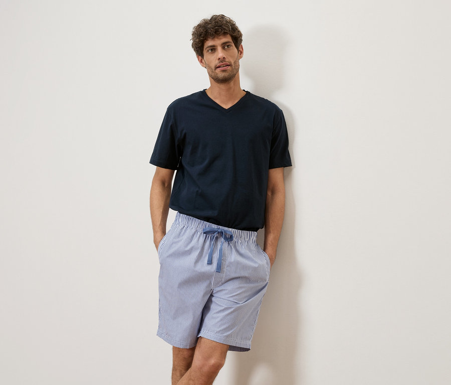 Un homme aux cheveux bouclés porte un t-shirt bleu marine et un short rayé, adossé à un mur blanc.