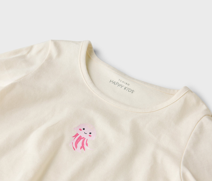 Gros plan d'un t-shirt crème avec un imprimé de méduse rose et l'inscription "Tchibo Happy Kids" au niveau du cou.