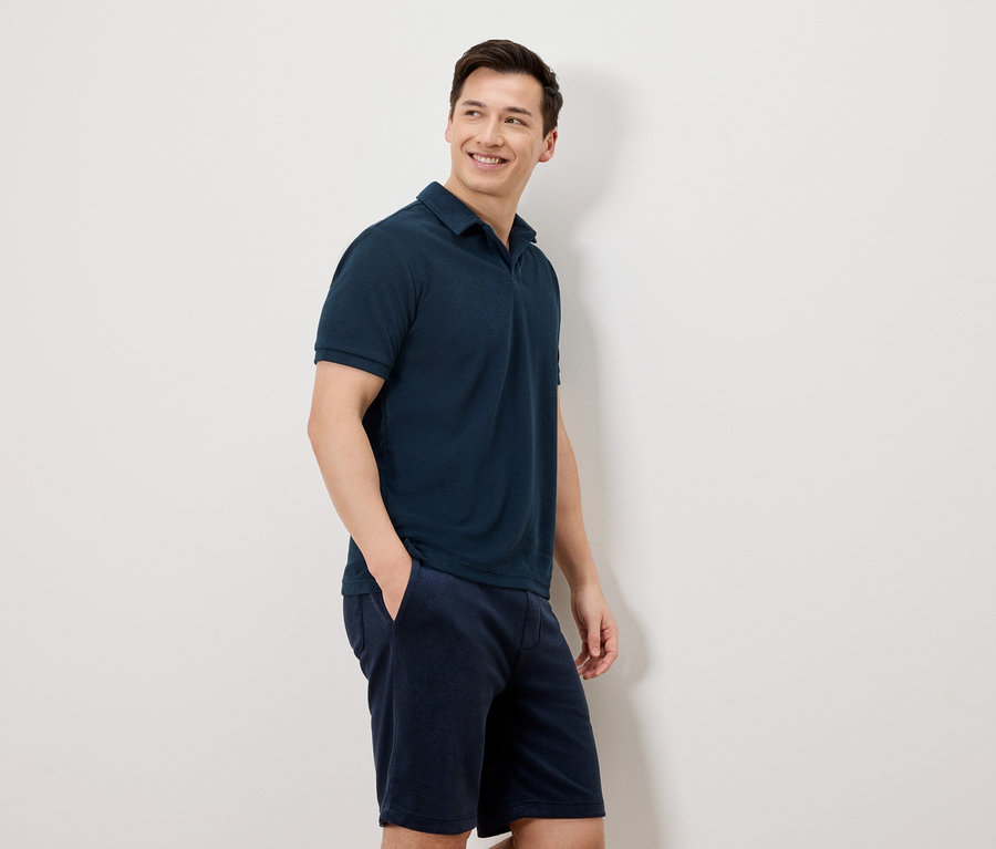 Un homme aux cheveux foncés portant un polo bleu marine et un short se tient devant un mur blanc.