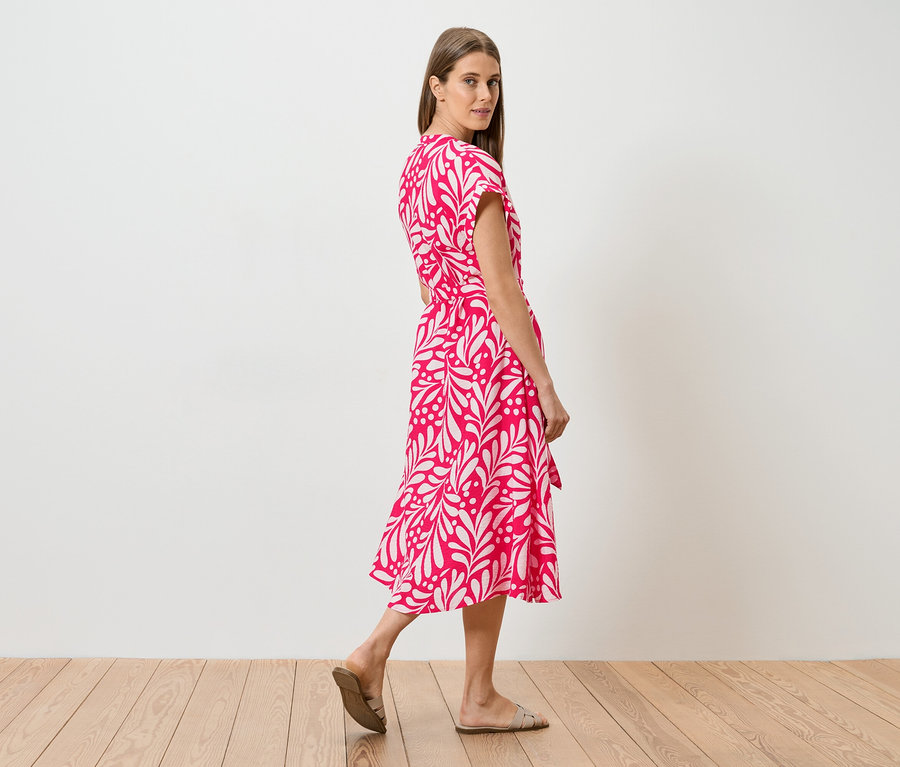 Une femme en robe rose à motifs de feuilles blanches marchant sur un plancher en bois, regardant par-dessus son épaule.