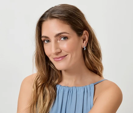 Une femme aux longs cheveux bruns ondulés et au haut bleu sourit à la caméra, portant des boucles d'oreilles bleues.
