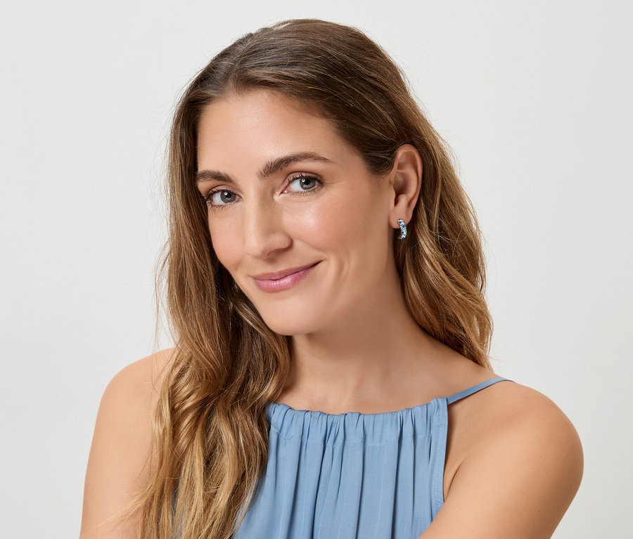 Une femme aux longs cheveux bruns ondulés et au haut bleu sourit à la caméra, portant des boucles d'oreilles bleues.