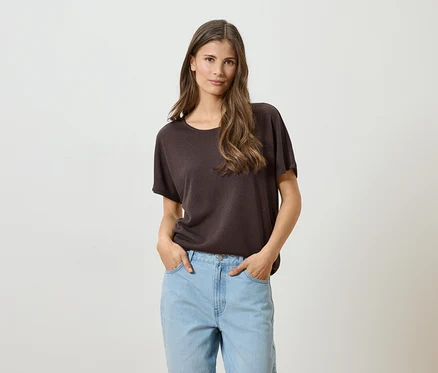 Une femme aux longs cheveux bruns, portant un t-shirt marron et un jean bleu clair, les mains dans les poches, sur un fond blanc.