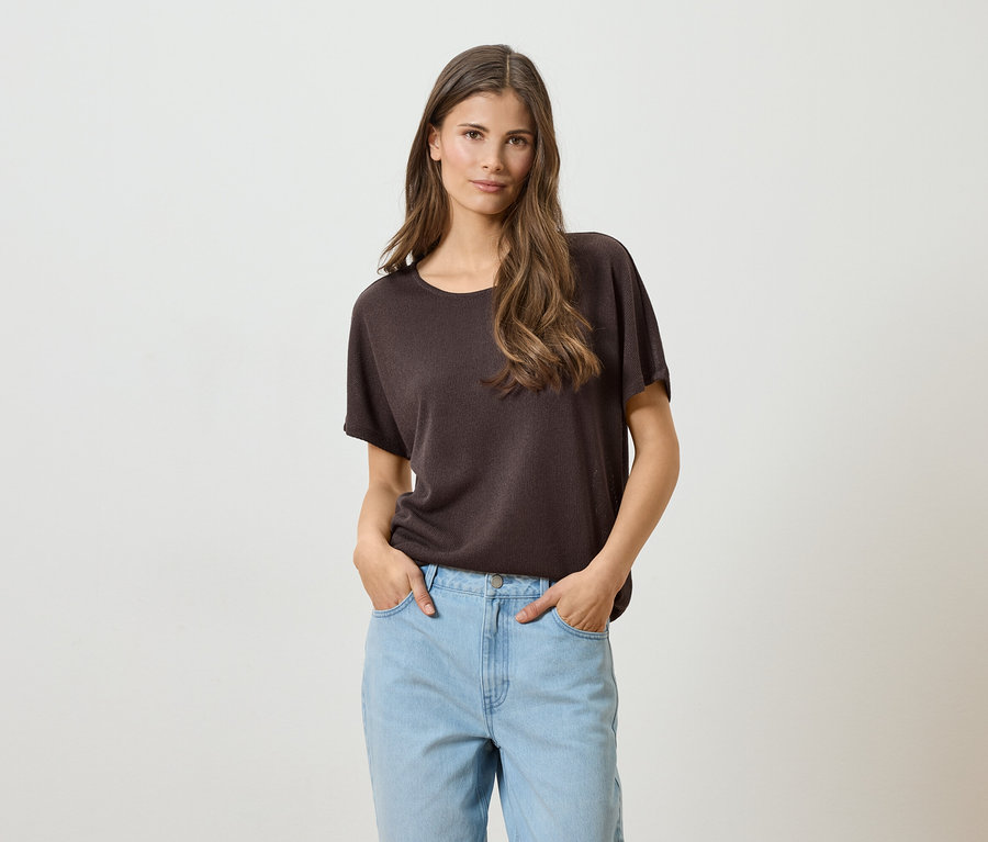 Une femme aux longs cheveux bruns, portant un t-shirt marron et un jean bleu clair, les mains dans les poches, sur un fond blanc.