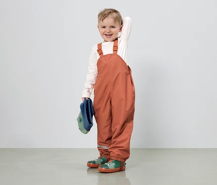 Un garçon souriant aux cheveux châtain clair porte une salopette orange, une chemise claire et des bottes en caoutchouc vertes à motifs d'animaux.