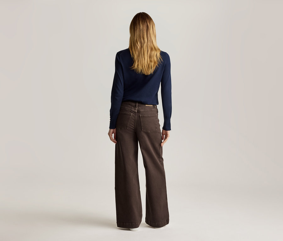 Vue de dos, une femme aux longs cheveux blonds porte un pull bleu marine et un pantalon marron ample.