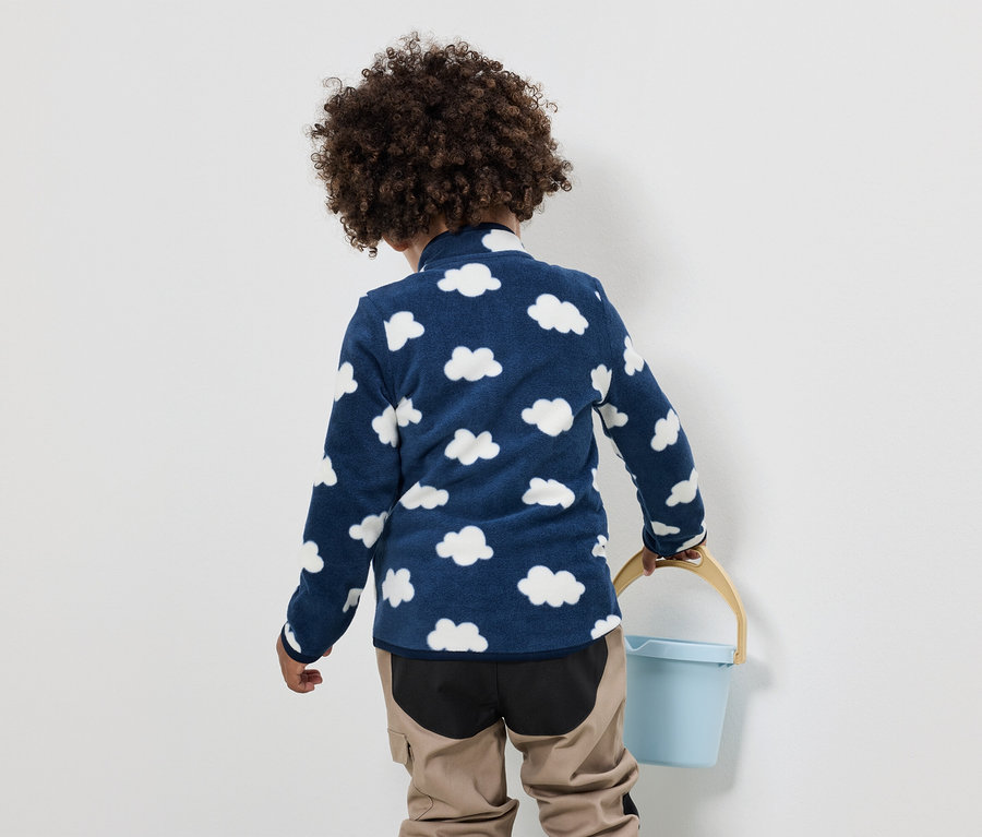 Un enfant aux cheveux bouclés porte un haut en polaire bleu avec un motif de nuages et un pantalon beige, et tient un seau bleu.
