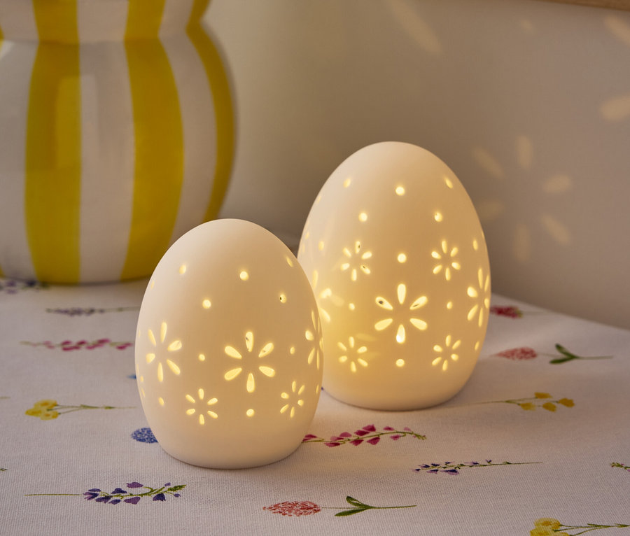 Deux lampes blanches en forme d'œuf avec des motifs floraux sont posées sur une table avec un vase de fleurs en arrière-plan.