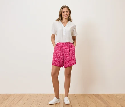 Une femme se tient devant un mur, les mains dans les poches. Elle porte un chemisier blanc, un short rose à motifs et des baskets blanches.
