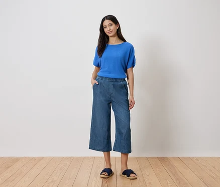 Femme debout, la main dans la poche, portant un haut bleu, une culotte de jeans et des sandales devant un mur blanc.