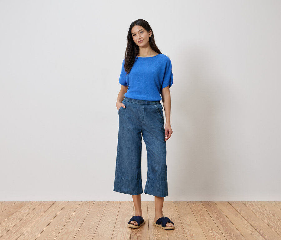 Femme debout, la main dans la poche, portant un haut bleu, une culotte de jeans et des sandales devant un mur blanc.