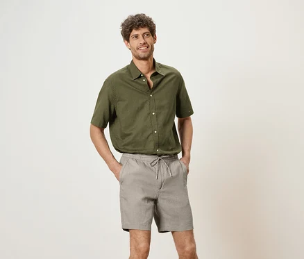 Homme portant une chemise vert olive et un short gris, les mains dans les poches, sur un fond neutre.