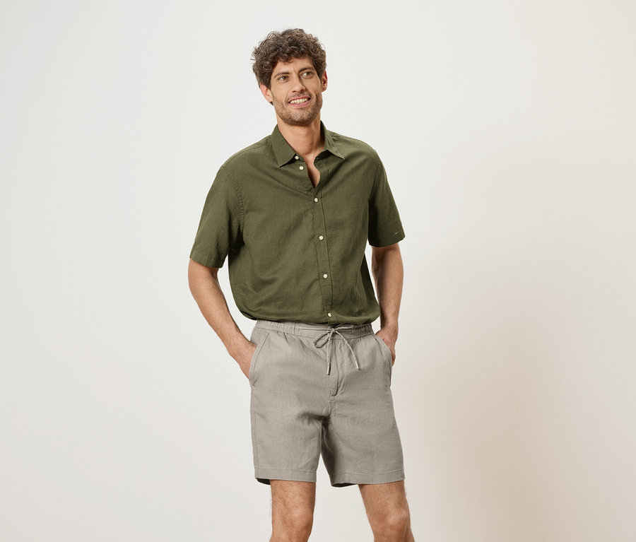 Homme portant une chemise vert olive et un short gris, les mains dans les poches, sur un fond neutre.