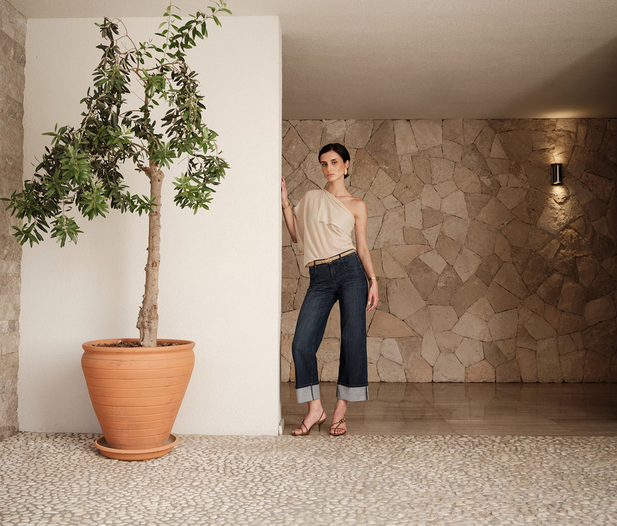 Une femme en jean et haut beige pose à côté d'un arbre en pot dans un intérieur aux murs en pierre.