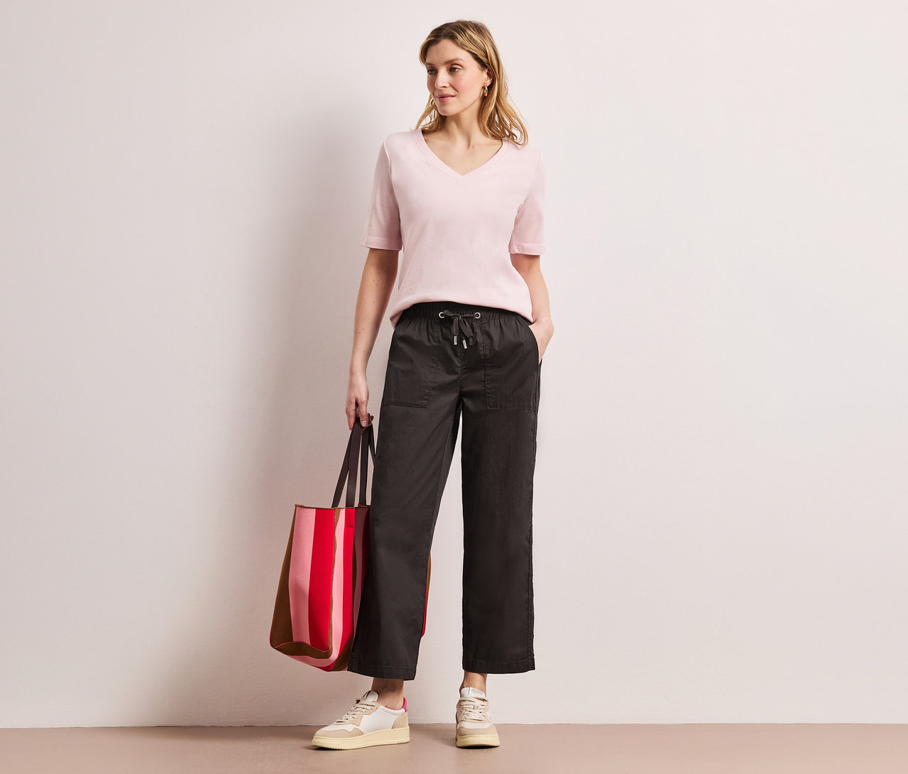 Une femme portant un haut rose et un pantalon Street One Papertouch pose avec un sac rayé rouge.