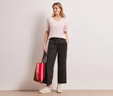 Une femme portant un haut rose et un pantalon Street One Papertouch pose avec un sac rayé rouge.
