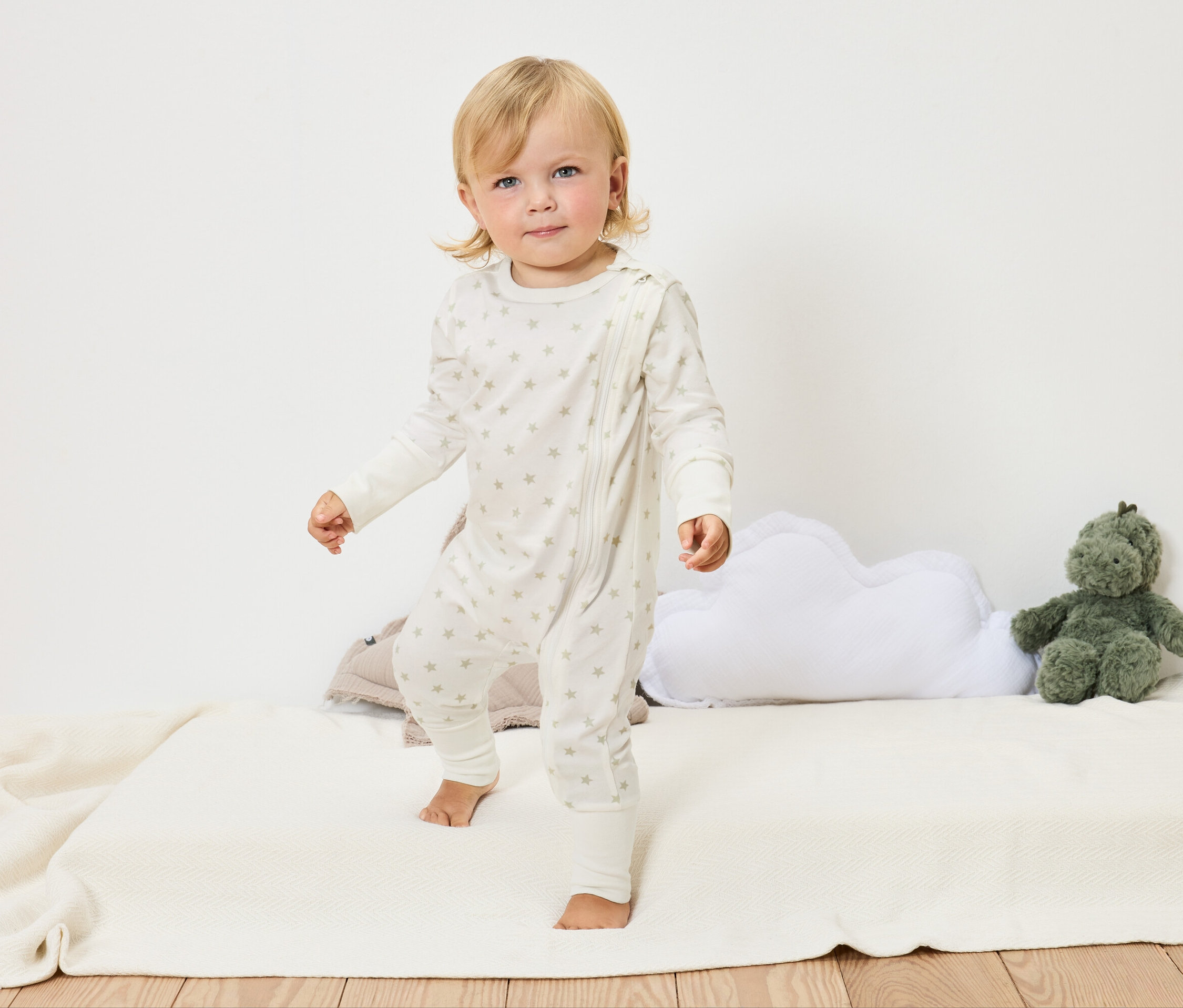 Un bébé blond en combinaison blanche à étoiles se tient pieds nus sur une couverture blanche à côté d'un coussin nuage et d'une peluche verte.