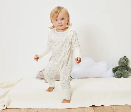 Un bébé blond en combinaison blanche à étoiles se tient pieds nus sur une couverture blanche à côté d'un coussin nuage et d'une peluche verte.