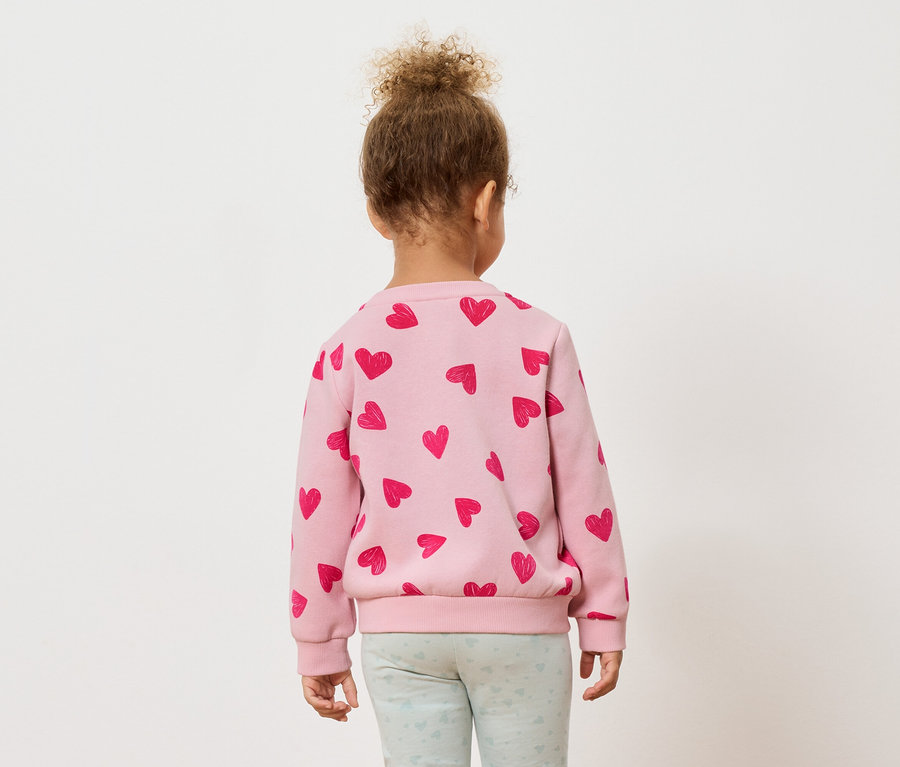 Vue de dos d'un jeune enfant aux cheveux bouclés, portant un pull rose avec des cœurs rouges et un legging bleu clair.