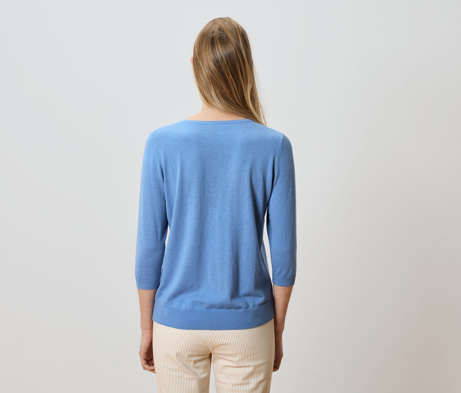 Vue arrière d'une femme aux longs cheveux blonds, vêtue d'un pull bleu clair et d'un pantalon rayé.