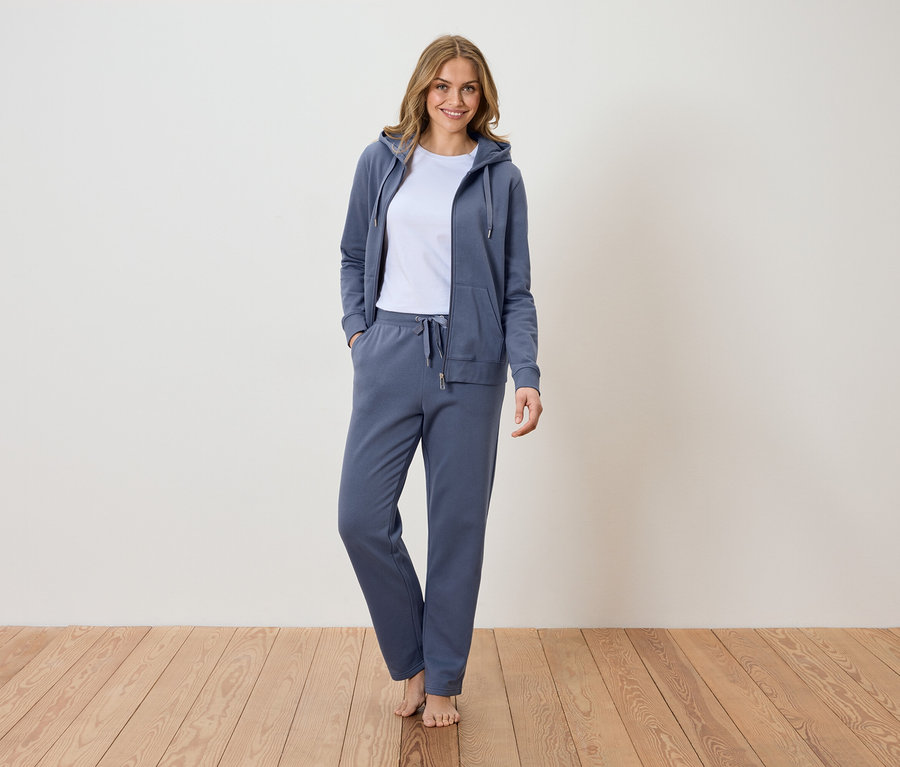 Une femme se tient pieds nus sur un plancher en bois, vêtue d'un survêtement bleu avec une veste à capuche sur un t-shirt blanc.